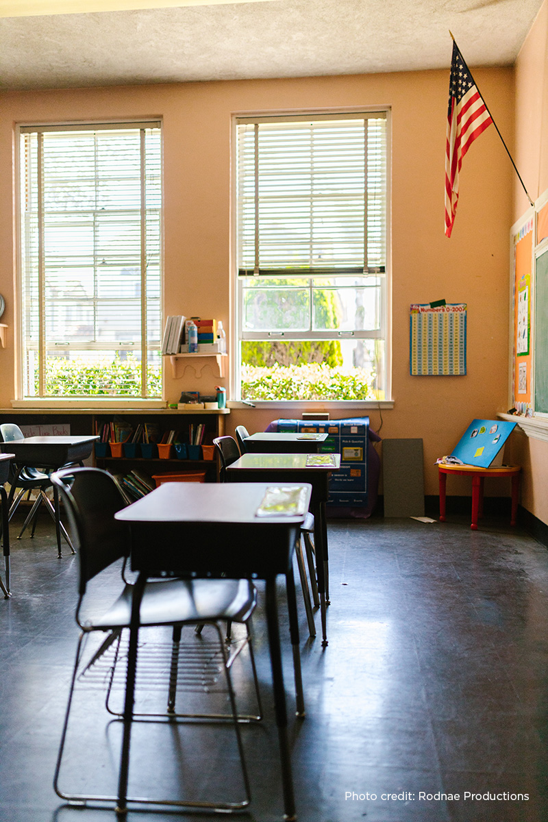 Elementary school classroom with desks and views of tress and shrubbery outside the classroom windows.